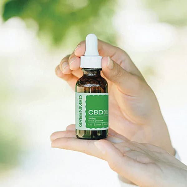 Mão segurando o frasco âmbar do óleo de CBD GreenMed CBDoil 2500 mg broad spectrum, com folhagem desfocada ao fundo